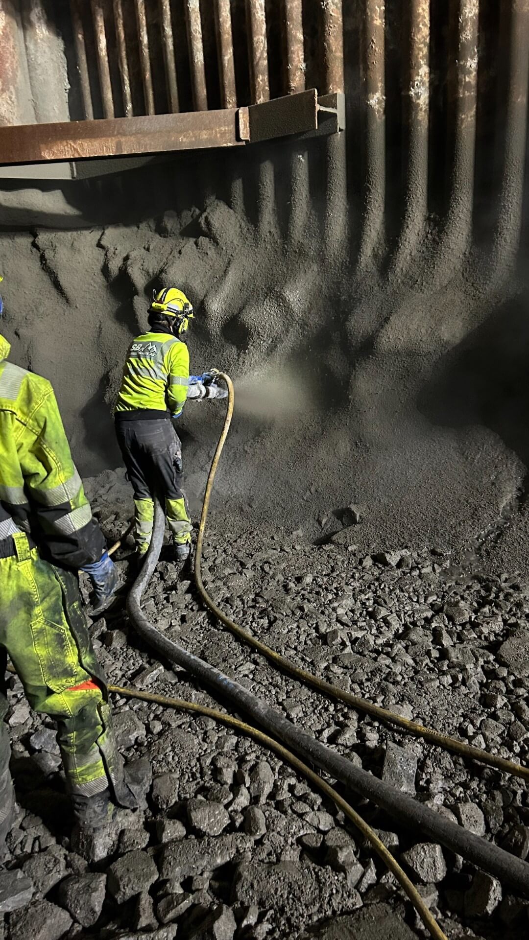 Arbeider utfører sprøytebetong på tunnelvegg med støtte fra kollega